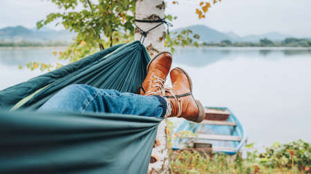 Young woman's leather fashion boots photo. Female swinging in a hammock between the birch trees on the mountain lake bank. Out-of-town Outdoor Recreation in Nature concept image.の写真素材
