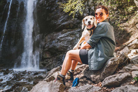 Smiling Female dog owner hugging his friend beagle dog resting near the mountain river waterfall during their together walking in autumn season time. Human and pets or walking in nature concept image.の写真素材