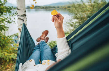 Young woman holding in fingers and examining small yellow birch tree leaf as she swinging in hammock on the mountain lake bank. Out-of-town Outdoor Recreation in Nature concept image.の写真素材