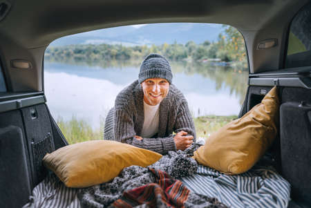 Sincerely smiling middle-aged man dressed in warm knitted clothes and jeans portrait in the cozy car trunk and with beautiful mountain lake view. Warm early autumn auto traveling concept image.の写真素材