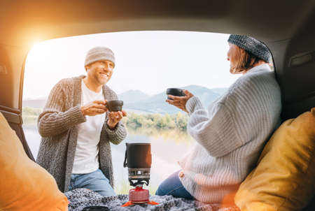 Car trunk view of chatting couple dressed warm knitted clothes enjoying gas stove prepared coffee and mountain lake view. Cozy early autumn couple auto traveling concept image.の写真素材