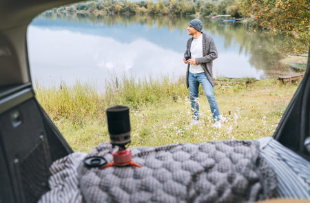 A man dressed warm knitted clothes enjoying gas stove prepared coffee and beautiful autumnal mountain lake view. Opened Car trunk shot. Warm early autumn auto traveling concept image.の写真素材