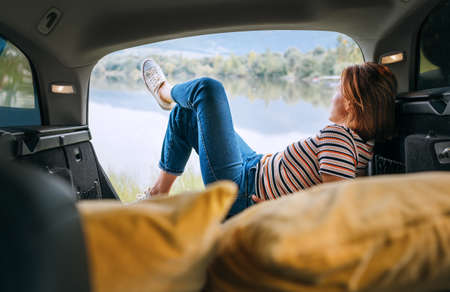 Woman dressed in warm calm colors clothes and jeans lying in the cozy car trunk and enjoying the mountain lake view. Warm early autumn auto traveling concept image.の写真素材