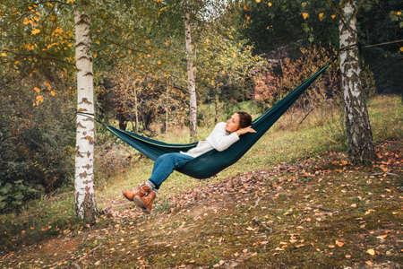 Young woman enjoying warm autumn weather and relaxing in swinging hammock between the birch trees....