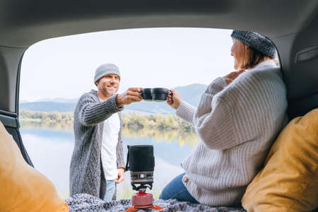Car trunk view clinking tea mugs couple dressed warm knitted clothes enjoying gas stove prepared drink and mountain lake view. Cozy early autumn couple auto traveling concept image.の写真素材