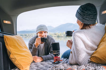 Car trunk view of chatting couple dressed warm knitted clothes enjoying gas stove prepared coffee and mountain lake view. Cozy early autumn couple auto traveling concept image.の写真素材