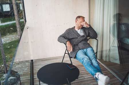 Portrait of a tired Middle-aged man dressed in open cardigan, jeans, and warm socks sitting on forest house balcony. He massaging tired eyes after. Vision health problems concept image.の写真素材
