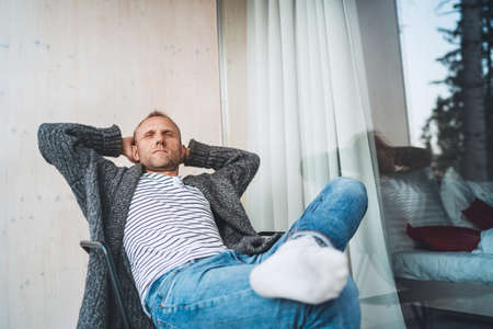 Portrait of a Middle-aged man dressed open cardigan, jeans, and warm socks sitting on forest house balcony and enjoying the fresh air. Everyday lifestyle concept image.の写真素材