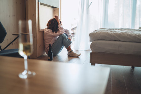 Sad woman sitting on the floor in the bedroom with a bottle of alcohol. Unfocused wine glass on the foreground table. Mental health and alcoholism problems concept image.の写真素材