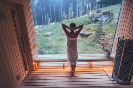Relaxing Woman wrapped white towel in Hot Finnish sauna with a huge wide window enjoying the green forest view and pleasant healthy body care temperature treatment. Body healthcare prevention conceptの写真素材