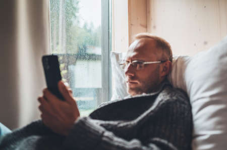 Window glass with raindrops photo with a middle-aged man dressed warm knitted cardigan lying on the cozy bed and using a modern smartphone. Everyday lifestyle photo with modern devices concept image.の写真素材