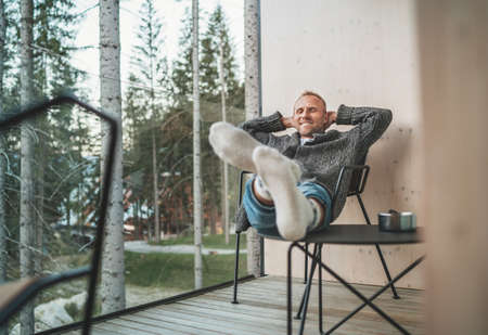 Portrait of a sincerely smiling Middle-aged man dressed open cardigan, jeans, and warm socks sitting in chair on forest house balcony and enjoying the fresh air. Everyday lifestyle concept image.の写真素材