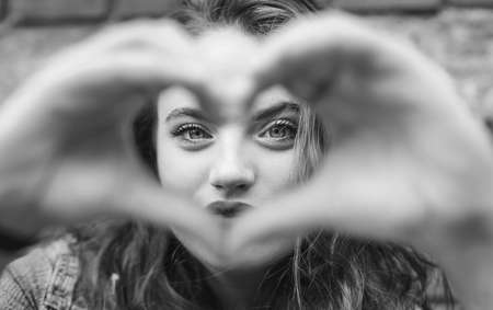 Beautiful Portrait of red curled long hair caucasian teen girl with applied red lipstick lips with blue eyes making a heart shape with fingers gesture. Love concept black and white image.の写真素材