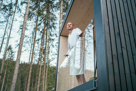 Smiling Man dressed white bathrobe standing on forest house balcony and enjoying fresh air with nature forest pine trees view. Outside Scandinavian interior design house. Living in wild concept imageの写真素材