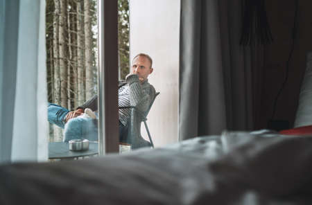 A middle-aged man dressed in open warm cardigan and jeans sitting on a forest house balcony and enjoying the fresh autumnal cold air. Everyday lifestyle concept from the room inside view image.の写真素材