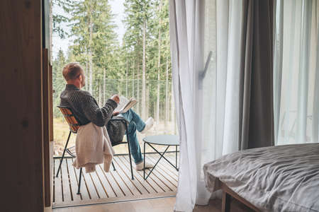 Alone man sitting in chair on country house balcony and enjoying forest view, tea cup, fresh air and bestseller novel thick book. Cozy bedroom interior back view. Reading or education concept imageの写真素材