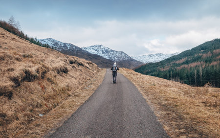 Lonely backpacker man walking with backpack by Scottish Highlands mountain road with snowy peaks at early moody morning. Active people in beautiful Nature concept image. Scotland, United Kingdom.の写真素材