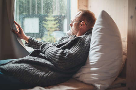 Portrait of thinking sad Middle-aged man in eyeglasses dressed open cardigan lying on cozy bed next...