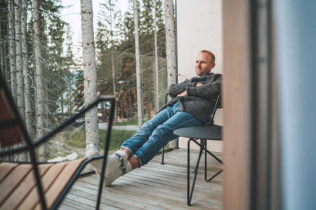 Portrait of a Middle-aged man dressed open cardigan, jeans, and warm socks sitting on forest house balcony and enjoying the fresh air. Everyday lifestyle concept image.の写真素材