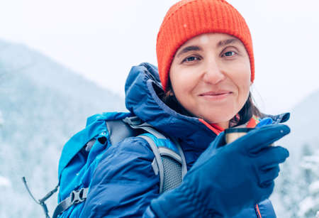 Middle-aged woman smiling, looking at camera drinking a hot drink from flask dressed warm down jacket while she trekking winter mountains route. Active people in the nature concept image.の写真素材