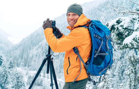 Smiling Man dressed orange softshell jacket with backpack photographing a landscape using digital camera and tripod while he trekking winter mountains route. Photographer occupation concept image.の写真素材