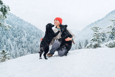 A young woman in warm clothes walking her 2 dogs in a picturesque snowy mountain outdoor. Female laughing and playing with pets and one dog licking an owner's cheek.Human and pets winter concept imageの写真素材