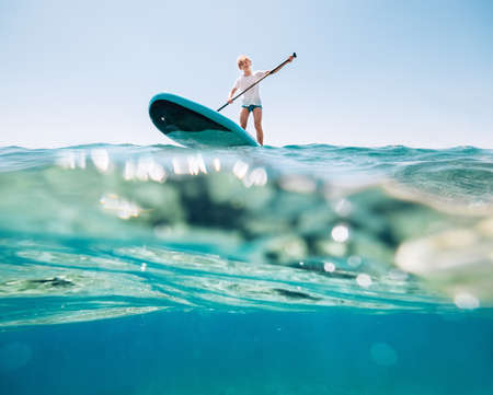Under the water view angle to the smiling blonde teenager, boy rowing stand up paddleboard. Active...