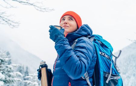 Middle-aged woman smiling and drinking a hot drink from flask dressed warm down jacket while she trekking winter mountains route. Active people in the nature concept image.の写真素材