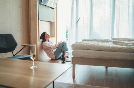 Sad woman sitting on the floor next to window in the bedroom with an opened bottle of alcohol. Unfocused white wine glass on the foreground table. Mental health and alcoholism problems concept image.の写真素材