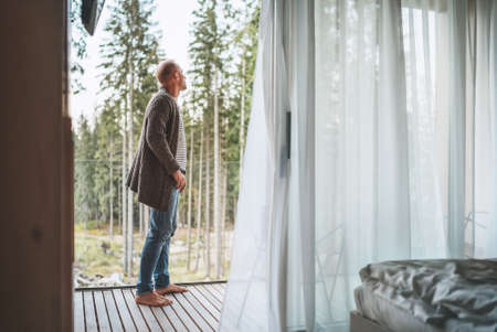 A man dressed open knitted cardigan standing on a forest house balcony and enjoying the fresh air with nature forest pine trees view. Outside Scandinavian interior design house. Living in wild conceptの写真素材