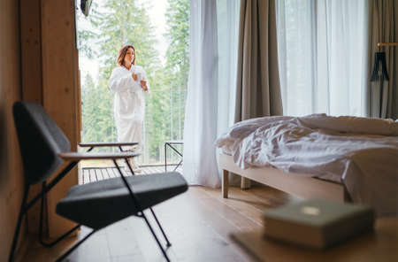 Woman dressed white bathrobe standing on forest house balcony and enjoying fresh air and morning teacup. Inside a Scandinavian interior design room view with a not made bed. Cozy living concept image.の写真素材