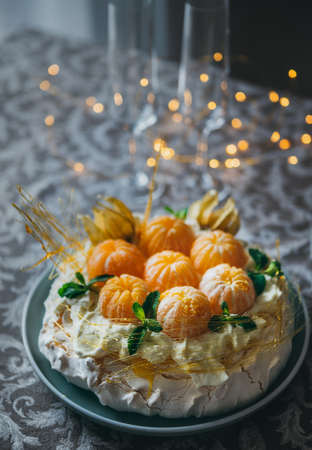 Pavlova cake is a meringue-based dessert named after the Russian ballerina decorated with fresh mandarines, caramel, mint, physalis, and powdered sugar with Christmas lights in background.の写真素材