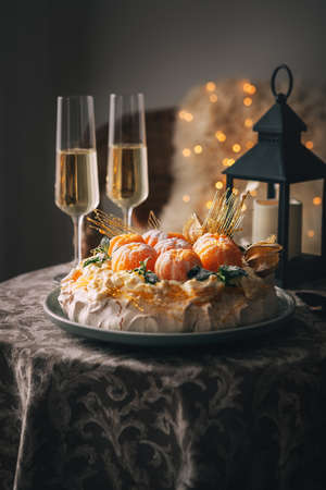 Pavlova cake is a meringue-based dessert named after the Russian ballerina decorated with fresh mandarins, caramel, mint, physalis, and powdered sugar with two champagne glasses on small table still.の写真素材