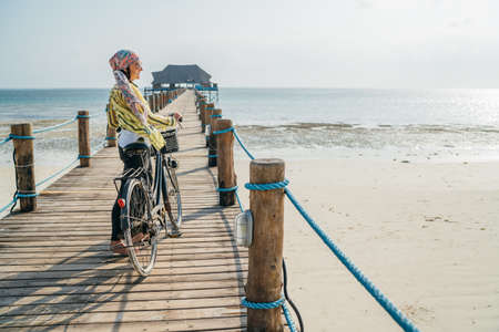 Portrait of a smiling woman dressed in light summer clothes, sunglasses with bicycle on the wooden sea pier on the sandy Zanzibar beach.Careless vacation in the tropical countries concept imageの写真素材