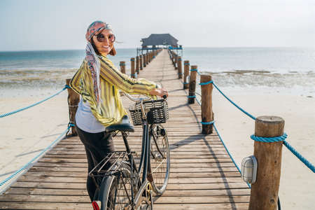 Woman looking at camera and smiling dressed in light summer clothes, sunglasses with bicycle on the wooden sea pier on sandy Zanzibar beach.Careless vacation in the tropical countries concept imageの写真素材