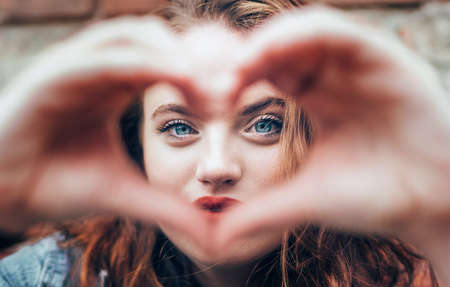 Beautiful Portrait of red curled long hair caucasian teen girl with applied red lipstick lips with blue eyes making a heart shape with fingers gesture. Love concept image.の写真素材