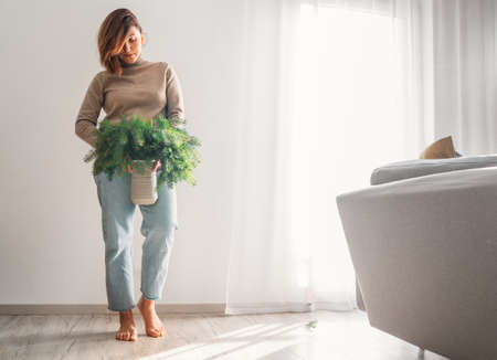 A young woman dressed warm cozy sweater holding the clay vase full of green freshly cut fir-tree coniferous branches near a white wall in the home living room with furniture. People and Nature conceptの写真素材