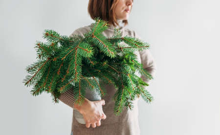 A woman dressed warm cozy sweater holding the clay vase full of green freshly cut fir-tree coniferous branches. Isolated on the white wall background people and Nature concept.の写真素材