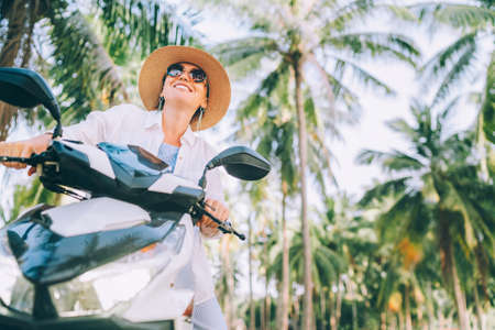 Dutch angle shoot of happy smiling woman in straw hat and sunglasses riding motorbike under palm...