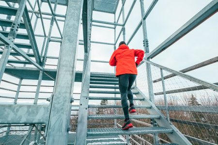Alone runner man dressed bright red softshell waterproof sporty clothes running up by huge steel industrial stairs on cold winter day. People healthy lifestyle concept photo.の写真素材