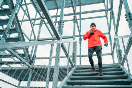 Smiling runner man dressed bright red softshell sporty clothes running down by huge steel industrial...