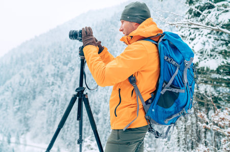 Photographer dressed orange softshell jacket with backpack making a landscape shoot using a digital camera and tripod while he trekking winter mountains route. Photographer occupation concept image.の写真素材