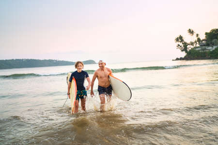 Portrait of a teen boy with his father with surfboards after surfing. They are smiling and walking...