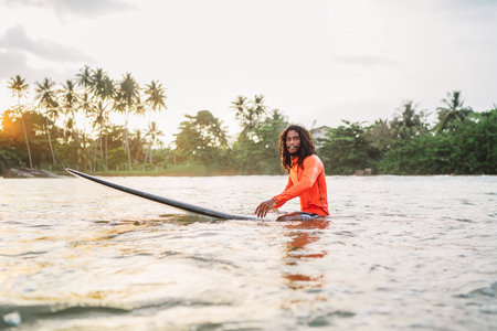 Black long-haired teen man floating on long surfboard, waiting for a wave ready for surfing with palm grove litted sunset rays. Extreme water sports and traveling to exotic countries concept.の写真素材