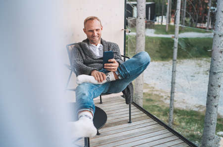 Smiling middle-aged man dressed open cardigan, jeans, and warm socks sitting on forest house balcony and using modern smartphone. Everyday lifestyle photo with modern devices concept image.の写真素材