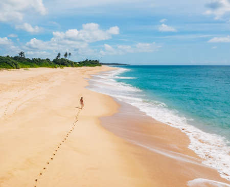 Young female dressed in light summer clothes walking barefoot leaving footprints on the sand on Indian ocean Tangalle lonely coconut trees beach on Sri Lanka island. Aerial top view drone shot.の写真素材