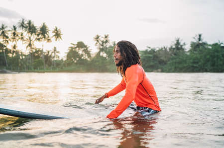 Black long-haired teen man floating on long surfboard, waiting for a wave ready for surfing with...