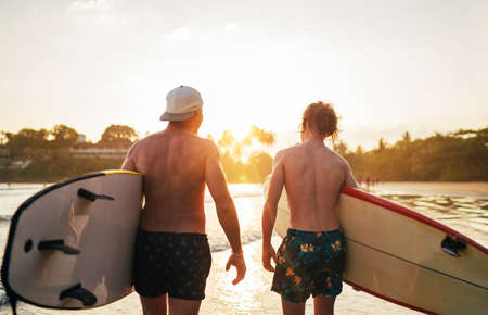 Father with teenager son walking with surfboards by the sandy ocean beach with palm trees on background lightened with sunset sun. They smiling and have a conversation. Family active vacation concept.の写真素材