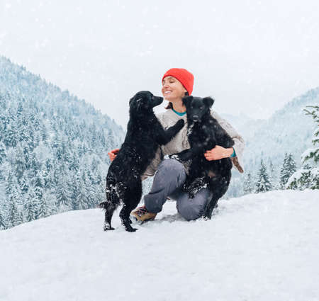 A young woman in warm clothes walking her 2 dogs in a picturesque snowy mountain outdoor. Female laughing and playing with pets and one dog licking an owner's cheek.Human and pets winter concept imageの写真素材