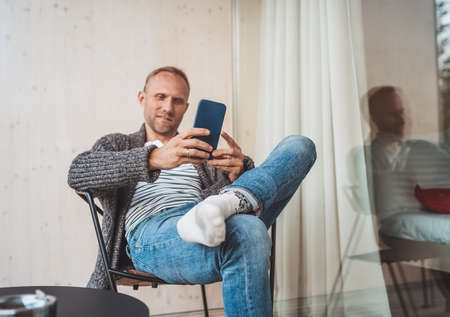 Smiling middle-aged man dressed open cardigan, jeans, and warm socks sitting on house balcony and smiling when he using modern smartphone. Everyday lifestyle photo with modern devices concept image..の写真素材